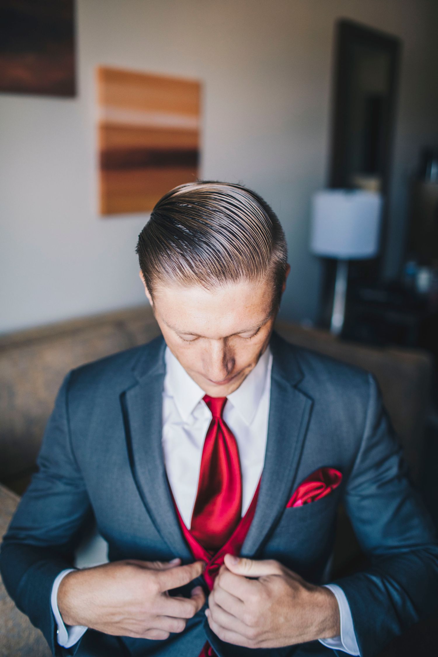 Groom in gray suit looking down while adjusting red tie and pocket square before wedding.