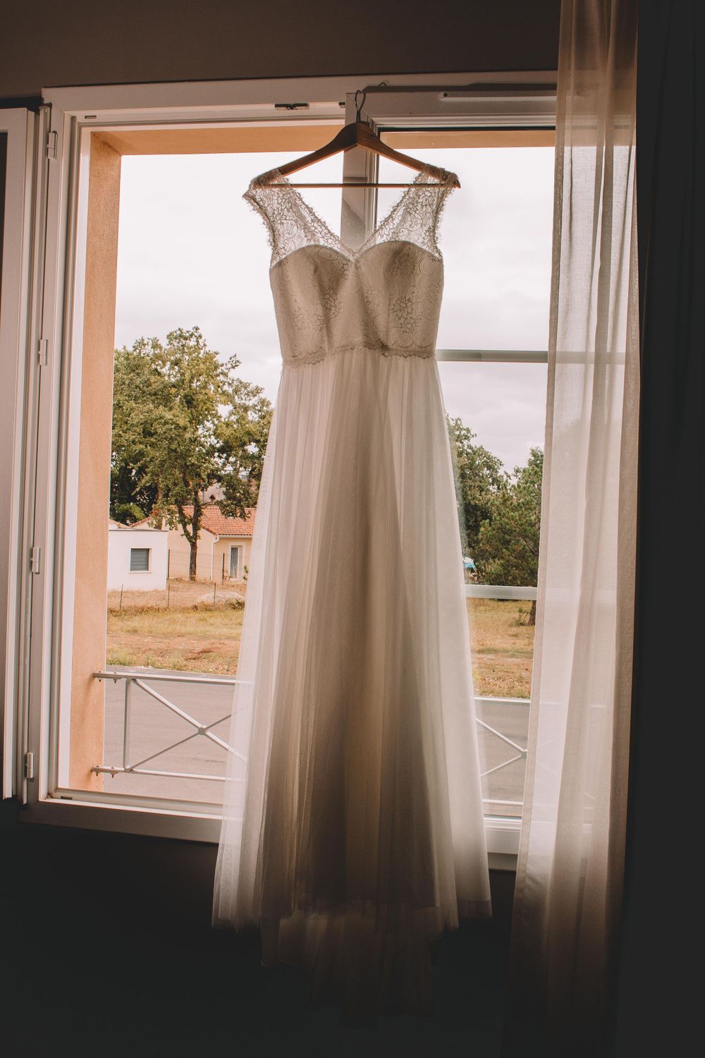 Elegant white wedding dress hanging in window with natural light streaming through sheer curtains.