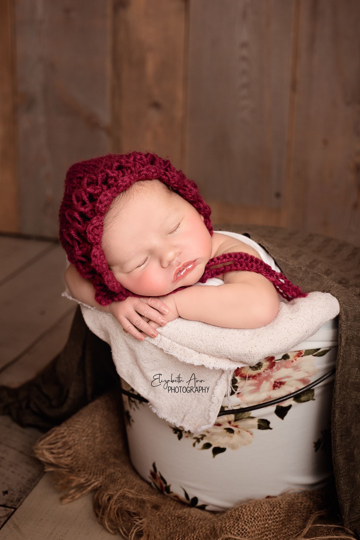 Newborn baby girl asleep in a floral bucket and wearing a bonnet, burlap and barn wood; Elizabeth Ann Photography WI
