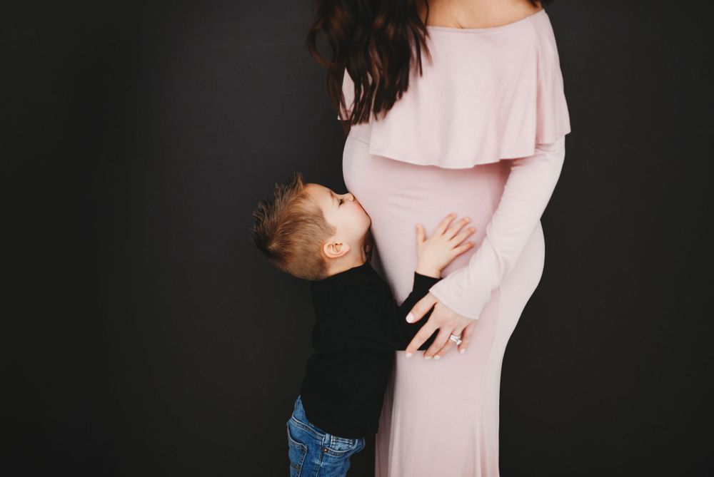 A young child in jeans hugs a pregnant woman wearing a soft pink off-shoulder dress against a dark background.