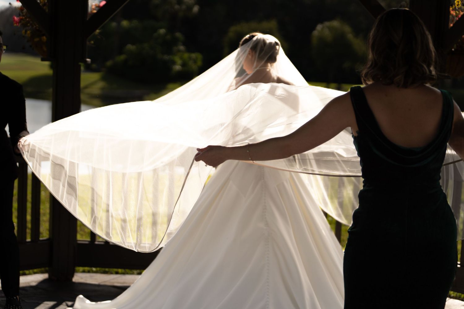 A flowing white wedding dress veil catches the sunlight as it spreads out with assistance from someone in a dark dress.