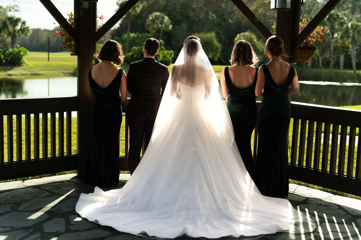 A bride wearing a white flowing gown stands with bridesmaids in black dresses on a covered deck overlooking a golf course.