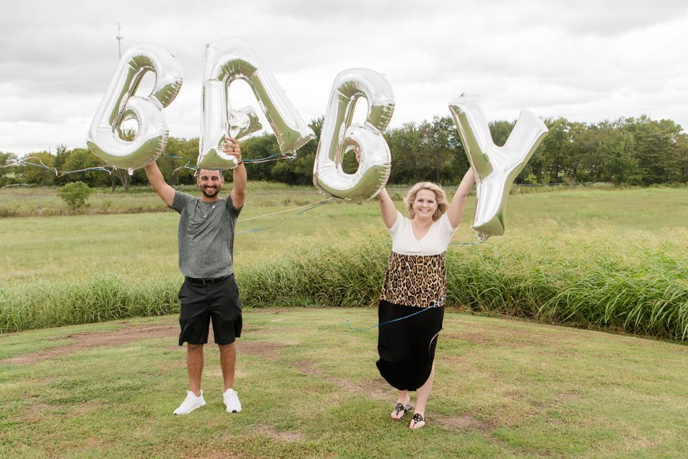 Couple holding silver BABY balloon letters overhead in field during pregnancy announcement photo shoot.