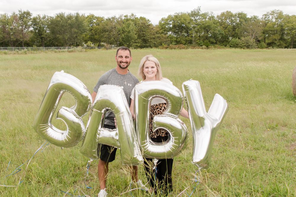 Couple stands in grassy field holding silver BABY balloons for pregnancy announcement photo.