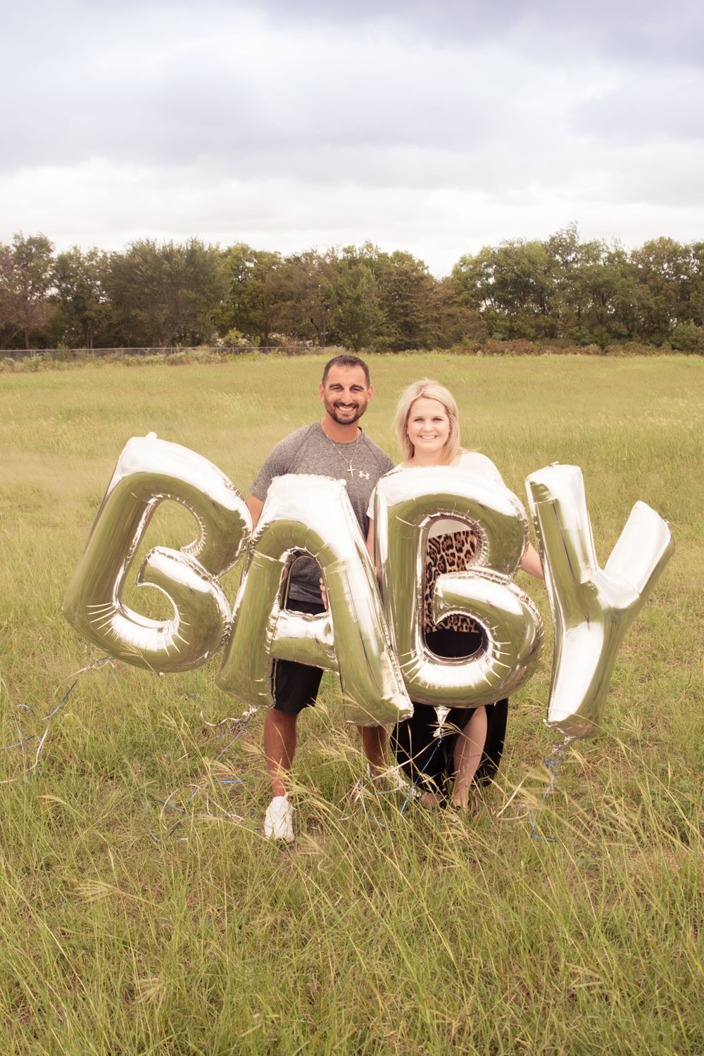 Couple poses with metallic BABY balloon letters in sunny field for pregnancy announcement.