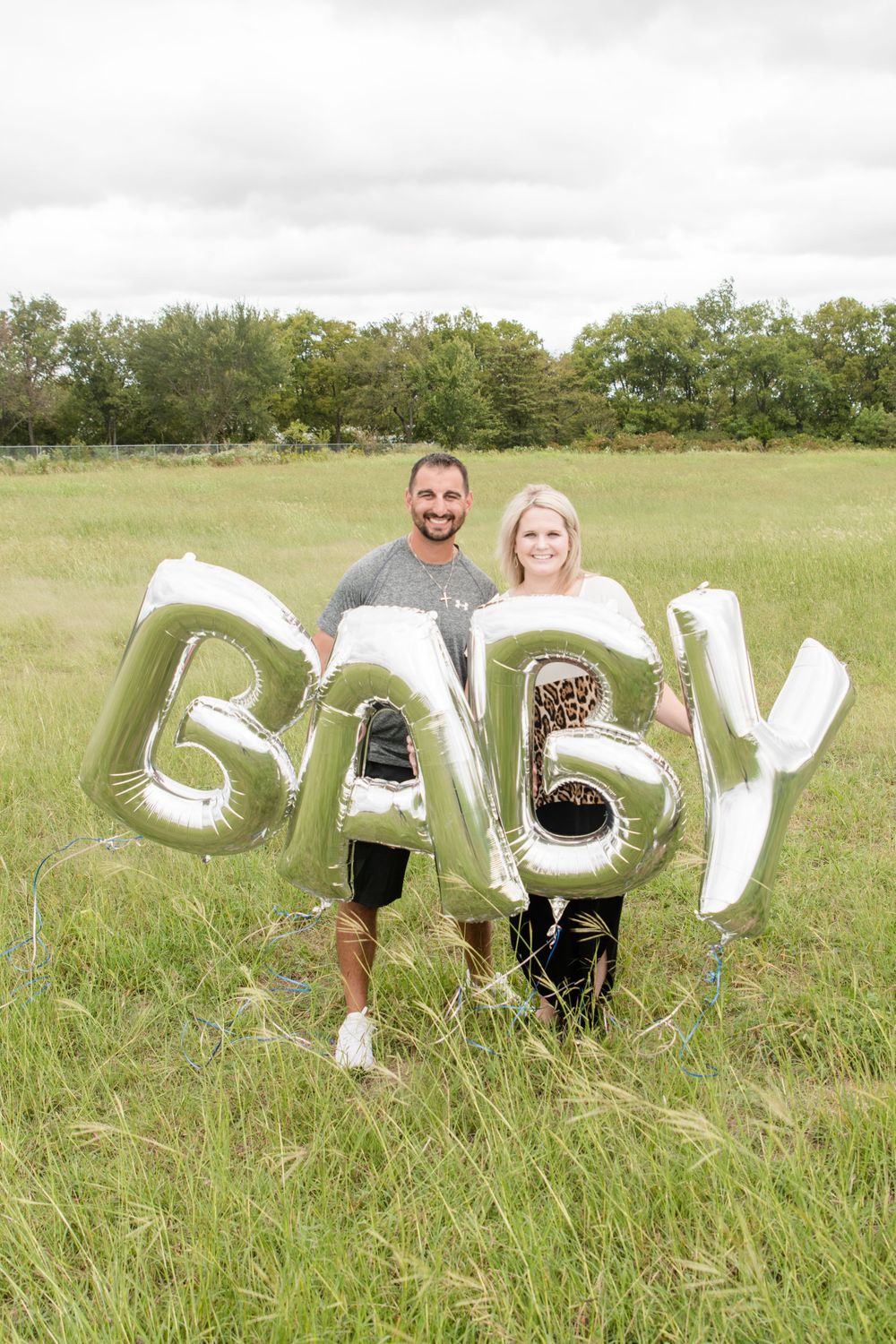 Couple holds large silver BABY balloon letters in field for maternity announcement shoot.