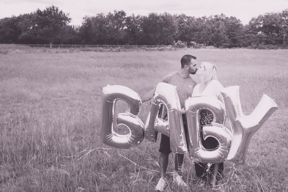 Black and white photo of couple handling silver BABY balloon letters in open field.