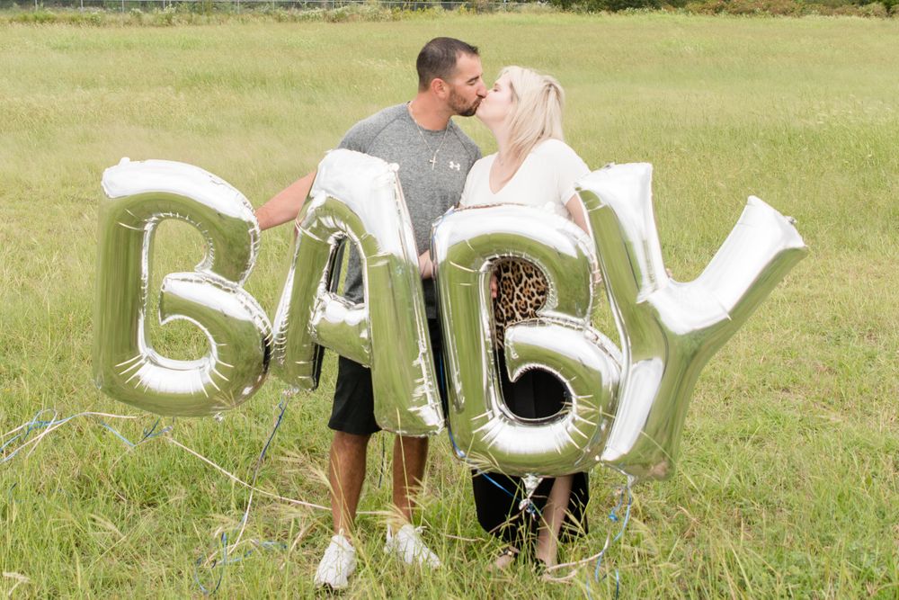 Couple poses with metallic BABY balloon letters during outdoor pregnancy announcement session.