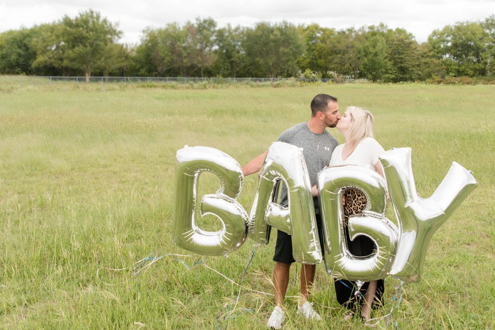 A couple stands in a grassy field holding silver BABY balloon letters for a pregnancy announcement photoshoot.