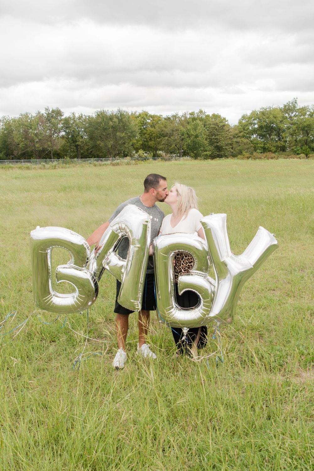 Couple sharing a romantic moment in a field while holding silver BABY balloons for their announcement photo.