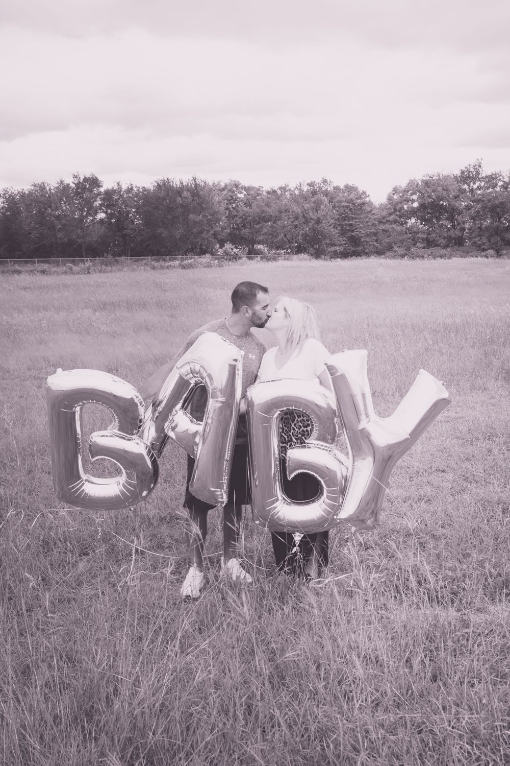 Black and white photo of couple embracing behind silver BABY letter balloons in an open meadow.