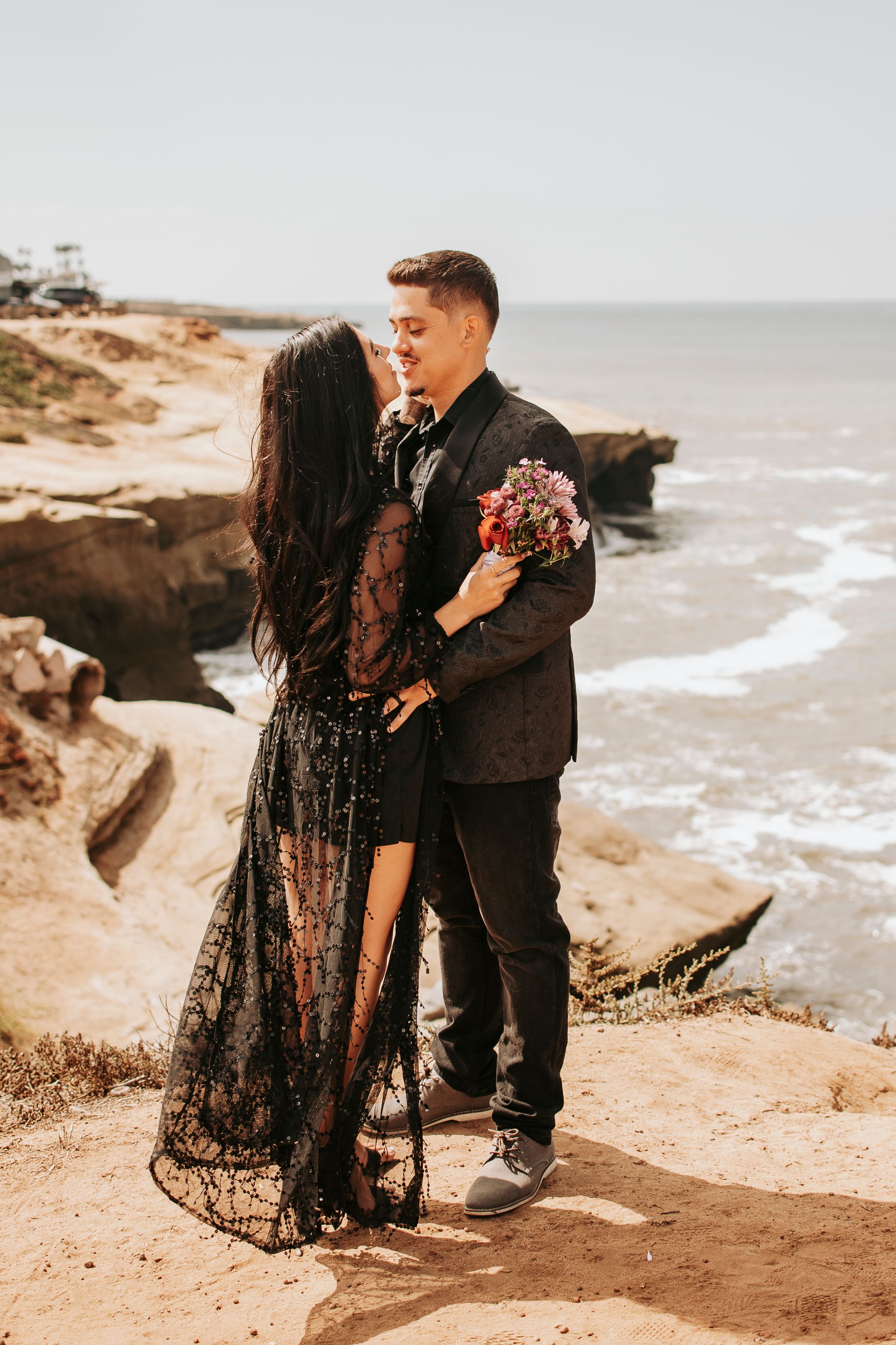 Couple in black attire posing romantically along rocky ocean coastline.