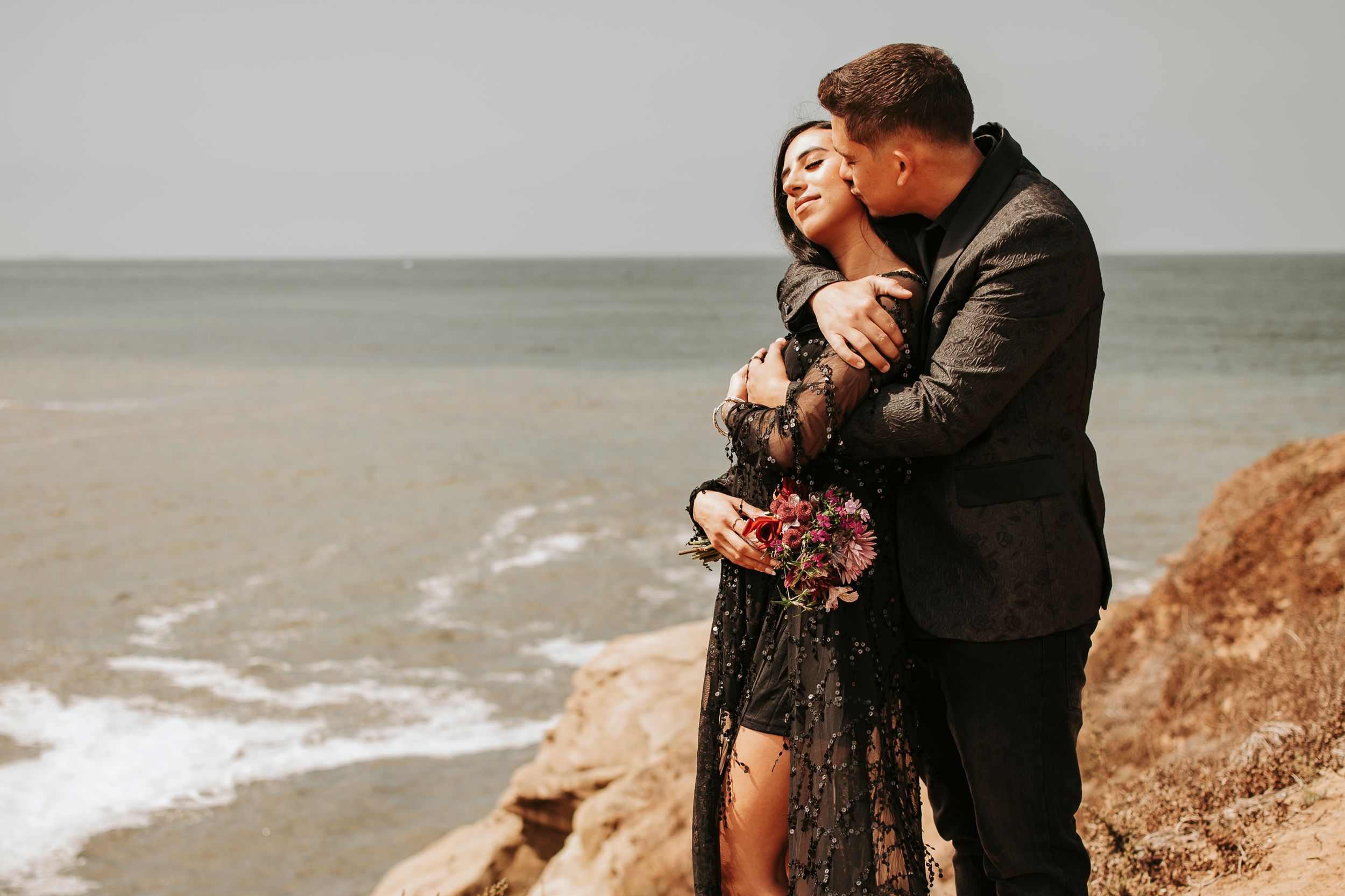 Romantic seaside photoshoot with couple embracing on coastal cliffs.