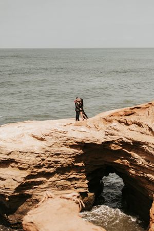 A couple stands intimately on a natural rock arch formation above the ocean.