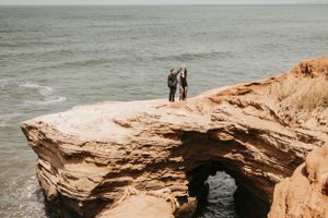 Two figures embrace on a dramatic coastal rock formation with an ocean arch beneath them.