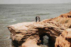 A couple stands together on a weathered sandstone arch overlooking the sea.