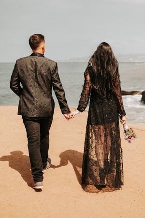 A couple in formal black attire holds hands while walking along a coastal path.
