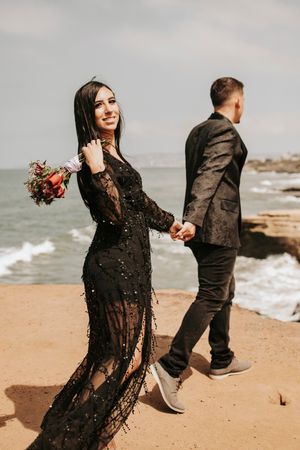 A couple in elegant black attire poses playfully on a beach with red flowers.