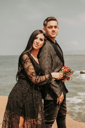 A couple in matching dark formal wear poses for a coastal portrait with red flowers.
