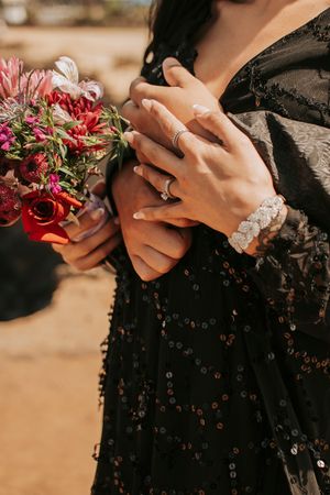 Close-up of hands holding red flowers showing engagement ring and sparkly black dress details.