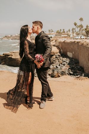 A romantic moment between a couple in black attire on a coastal cliff with palm trees.