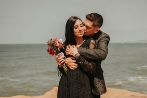 A tender embrace between a couple in formal wear holding red flowers by the ocean.