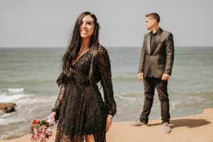 A couple poses on a beach with the woman in a black lace dress holding flowers while her partner stands behind.