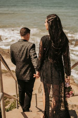 A couple descends stairs to a beach while holding hands, with the woman wearing a flowing black lace dress.