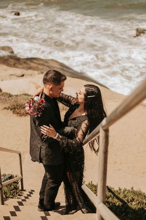 A romantic couple shares an intimate moment on beach stairs while holding a bouquet of pink flowers.