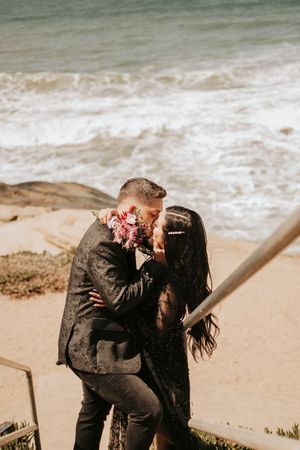 A couple embraces on coastal steps with ocean waves in the background and pink flowers between them.