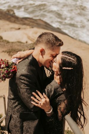 A couple shares a romantic moment on beach stairs while showing off an engagement ring.