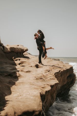 A couple shares a playful moment while standing on dramatic coastal rocks overlooking the ocean.