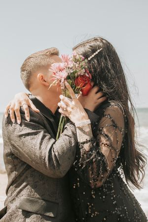 A couple shares a tender moment behind pink flowers on a cloudy beach day.