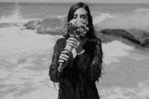 A person in a black lace dress holds a bouquet against ocean backdrop in dramatic black and white photography.