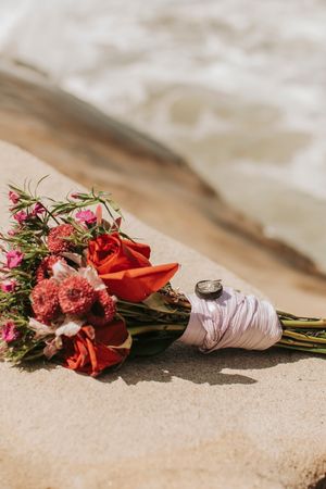 Wedding bouquet with red roses and pink flowers resting on sandy beach with ribbon wrapping.