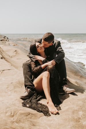 Couple in dark clothing sharing romantic moment on beach rocks.