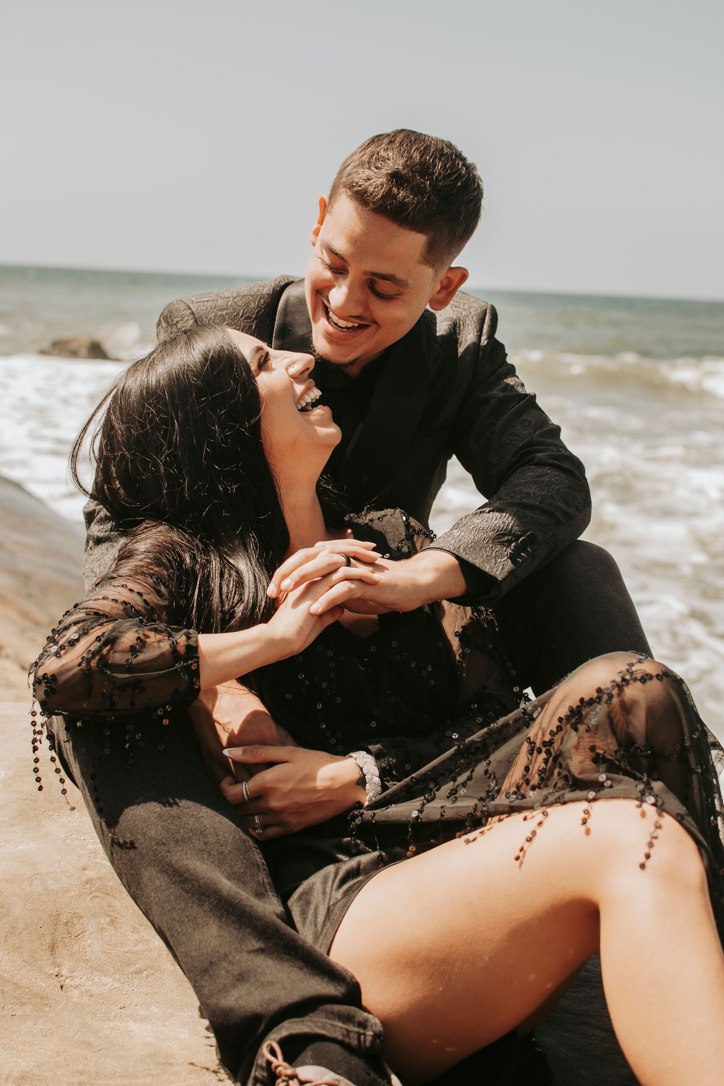 Playful couple in dark attire laughing together on sandy beach.