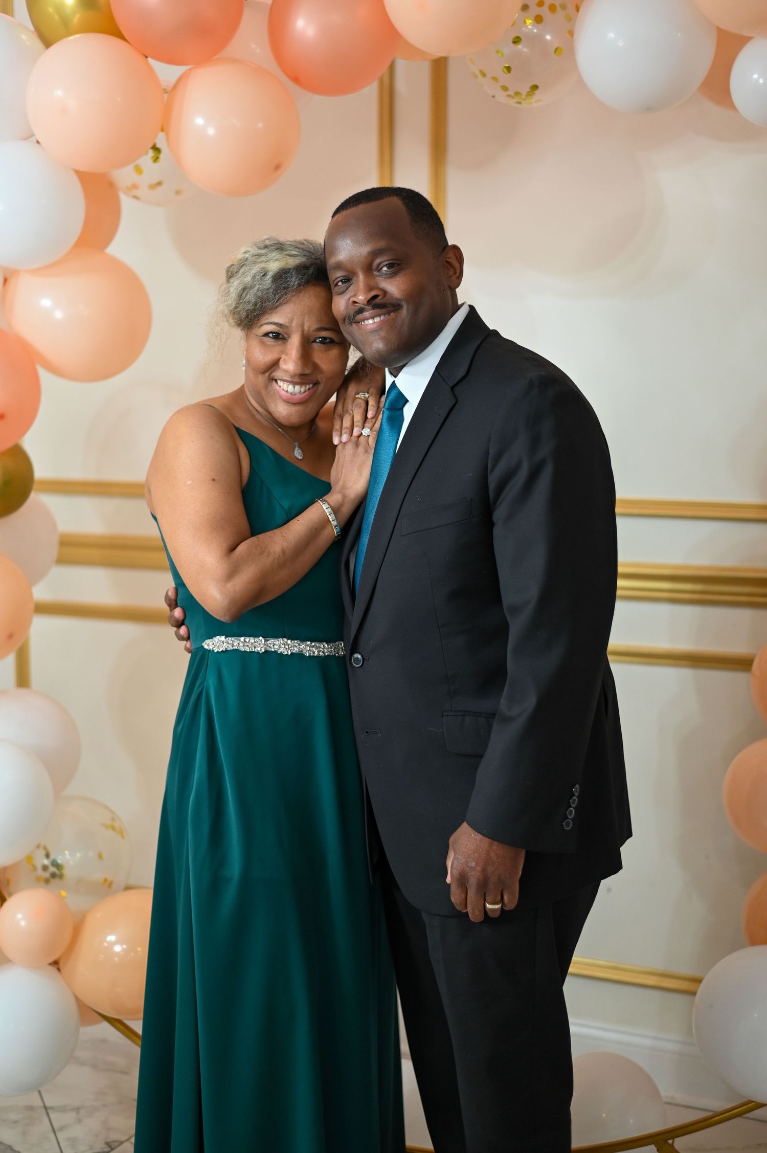 Formal couple in emerald gown and black suit posing under peach balloon arch at celebration.