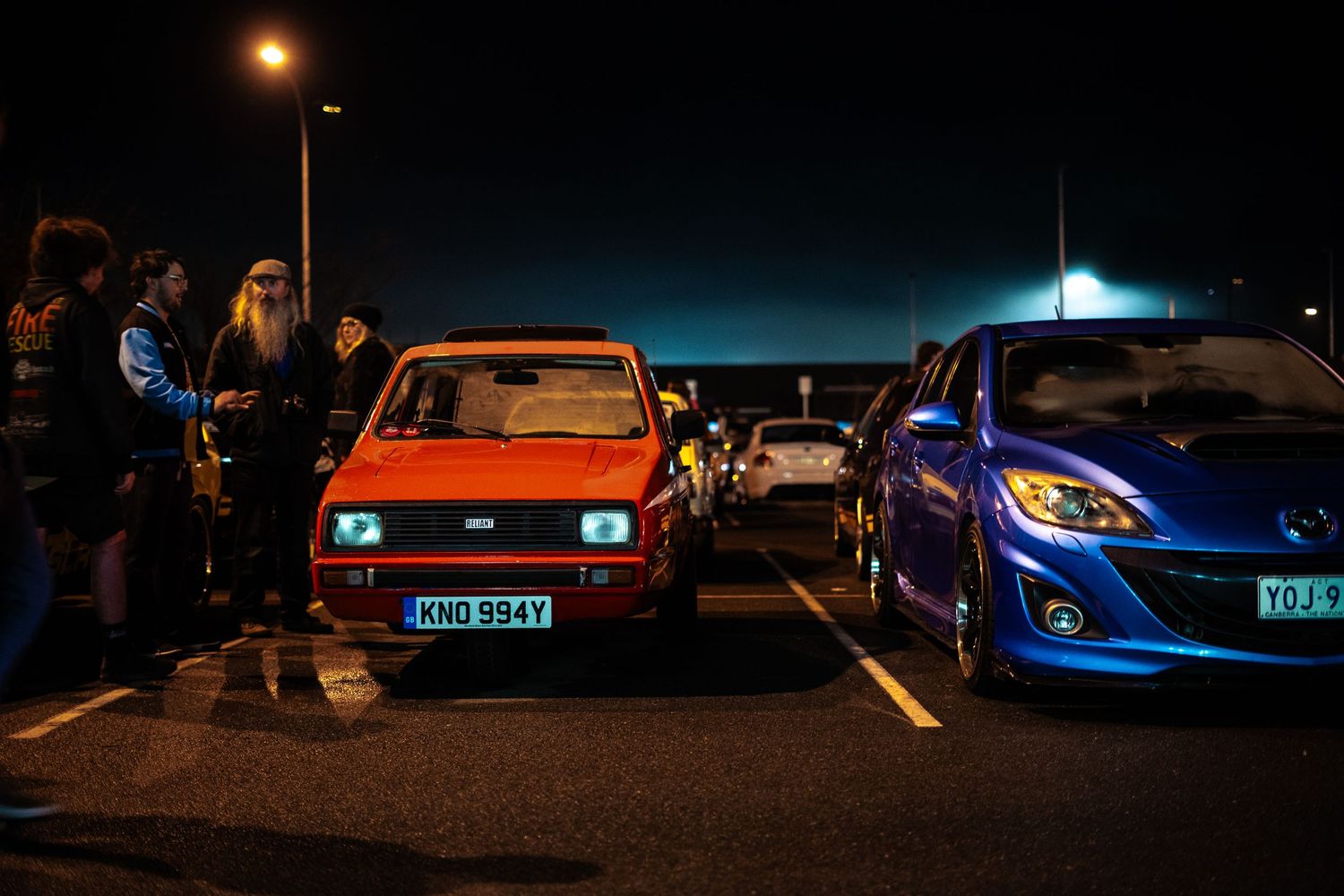 Vintage red hatchback parked next to modern blue sports car in nighttime parking lot.