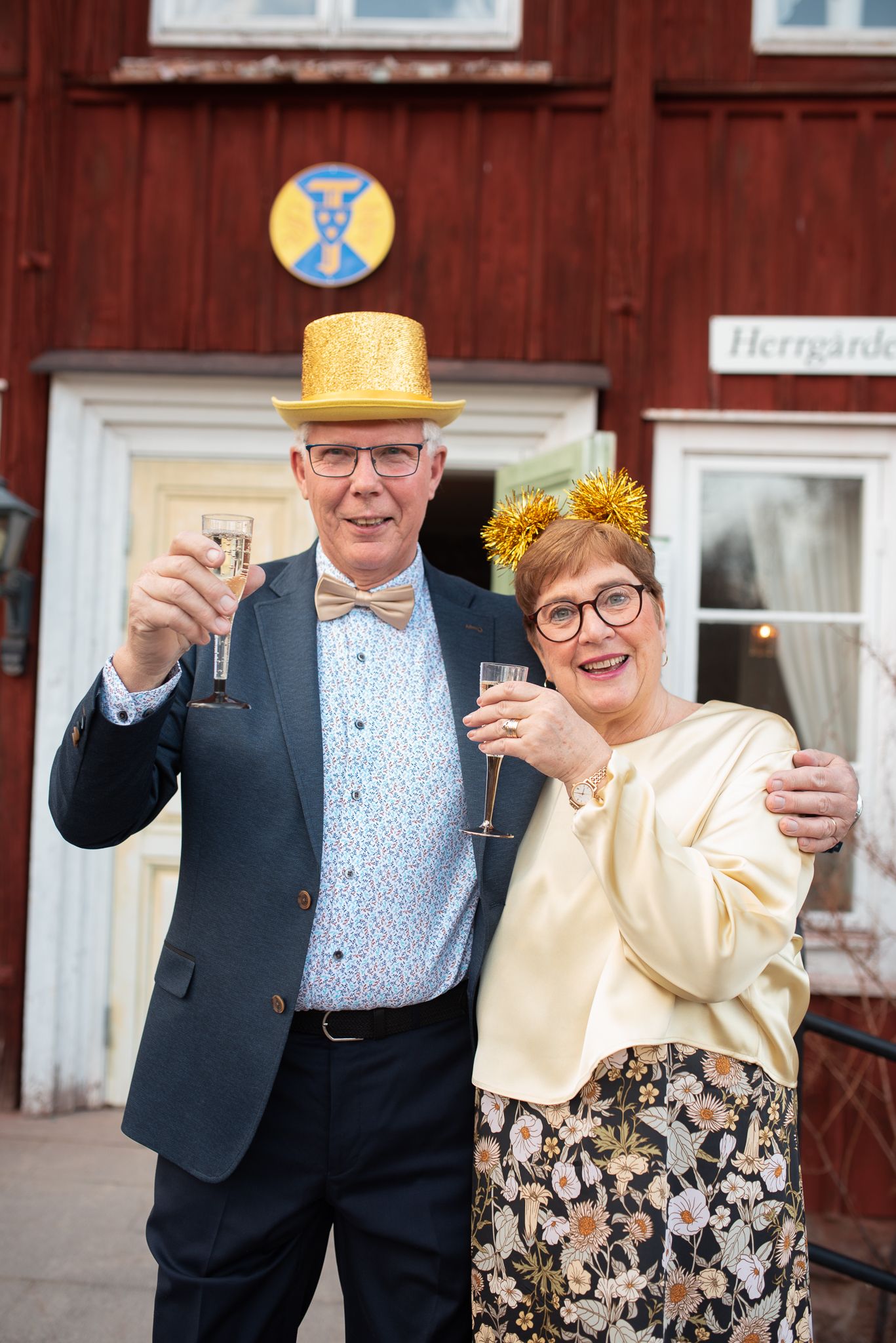 Festive celebration scene outside a red wooden building with people dressed in formal attire.