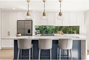Modern white kitchen with navy blue island, upholstered barstools, and glass pendant lights.