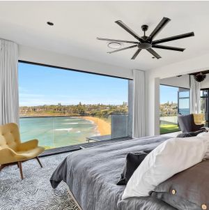 Modern bedroom with large windows overlooking beach coastline and oversized ceiling fan.