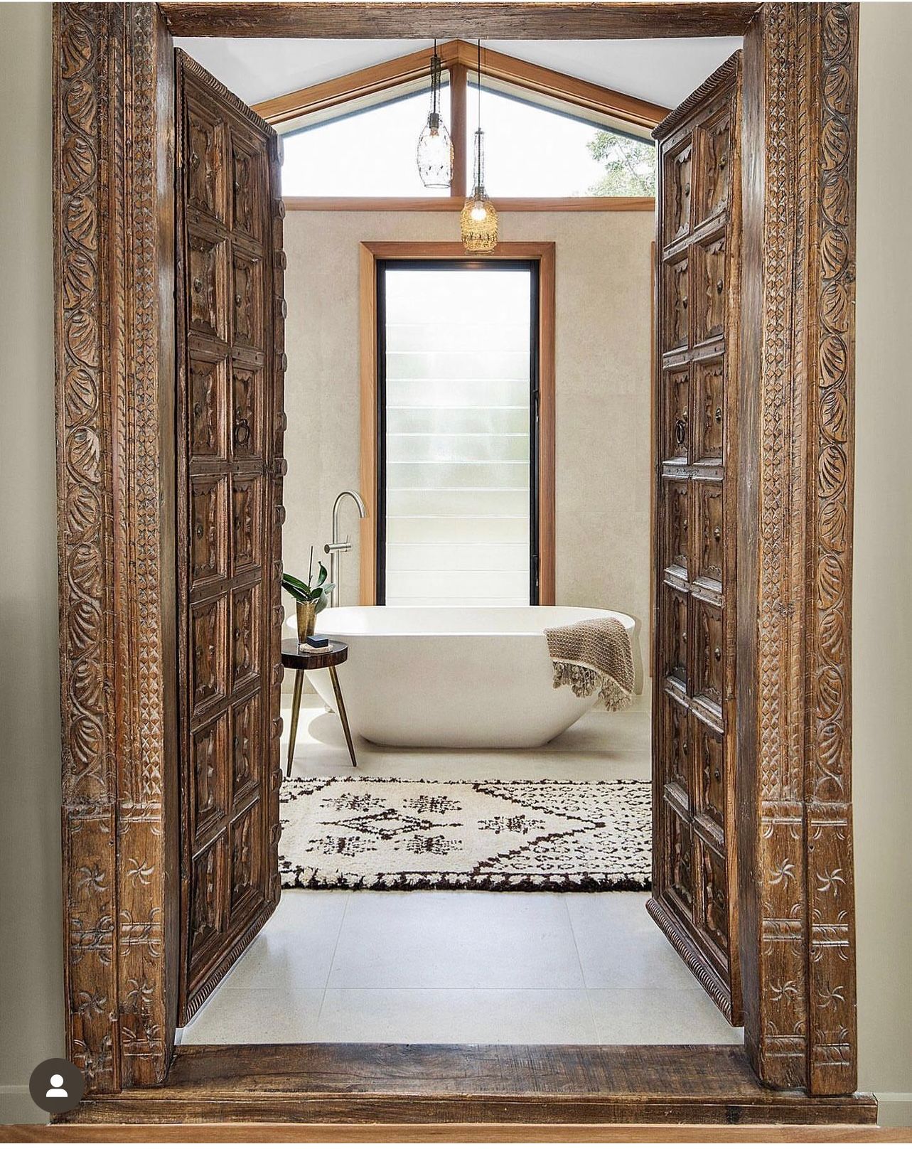 Ornate carved wooden double doors leading to bathroom with freestanding tub and patterned rug.