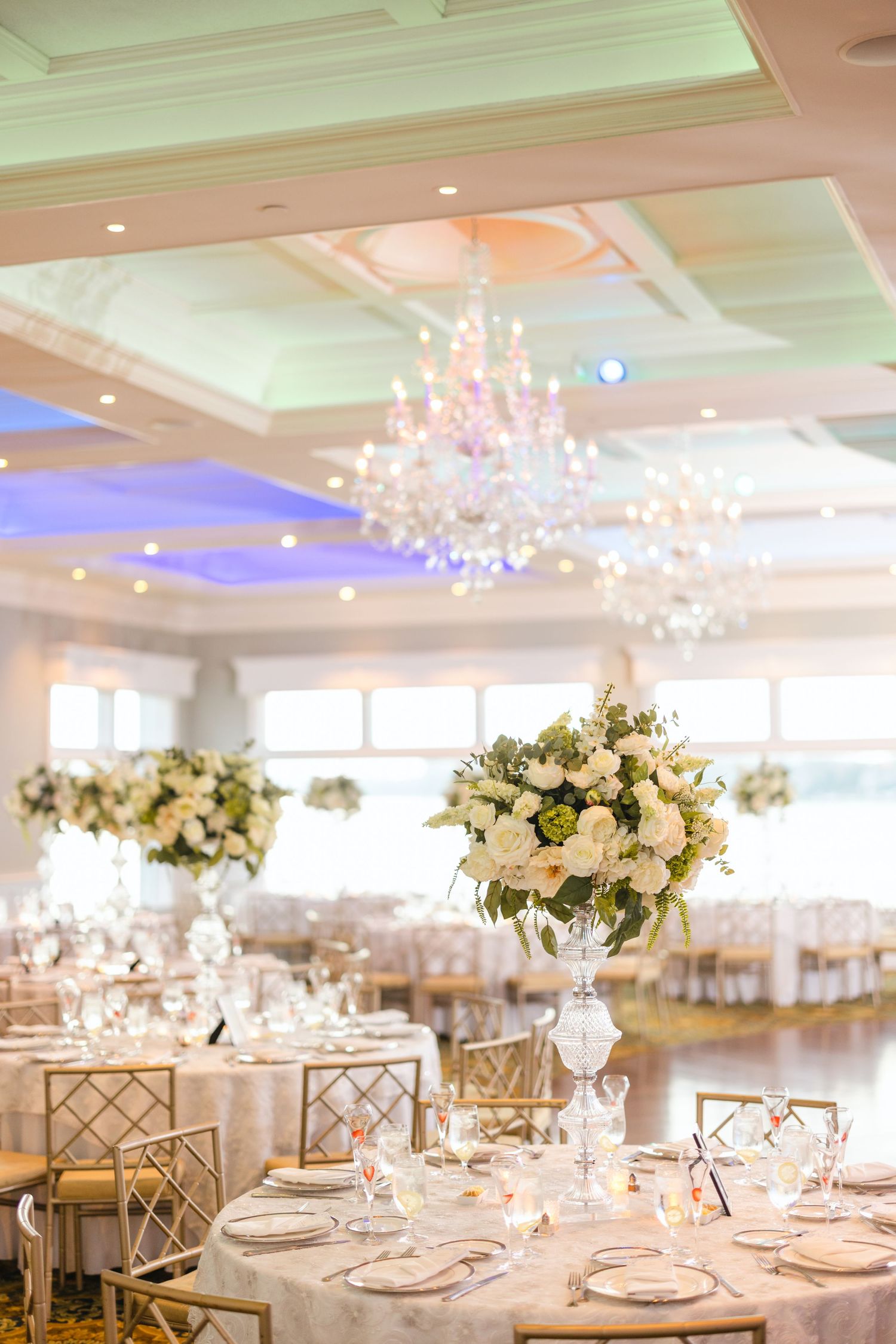 Elegant wedding reception ballroom with crystal chandeliers and tall white floral centerpieces on round tables.