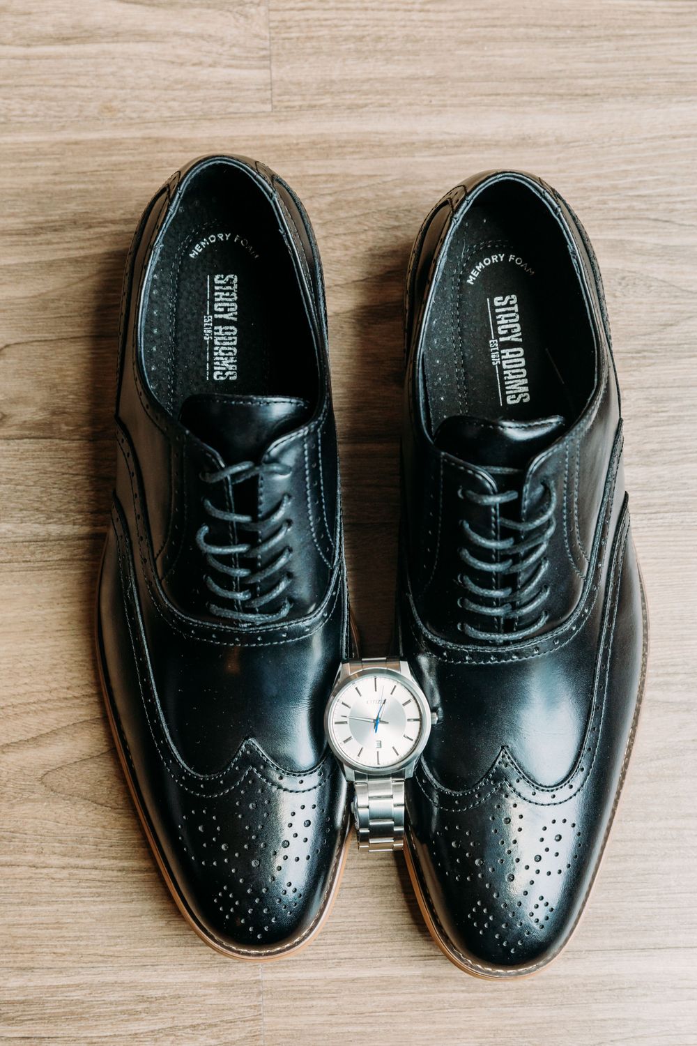 Black formal dress shoes and wristwatch arranged on wooden floor.