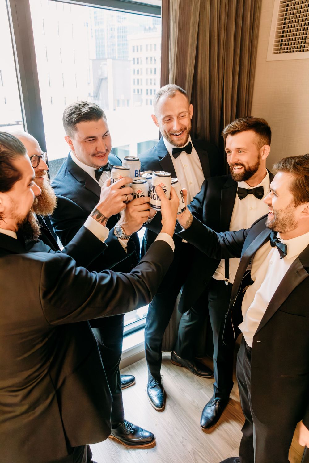 Groomsmen in black tuxedos and bowties sharing laughs in a hotel room.