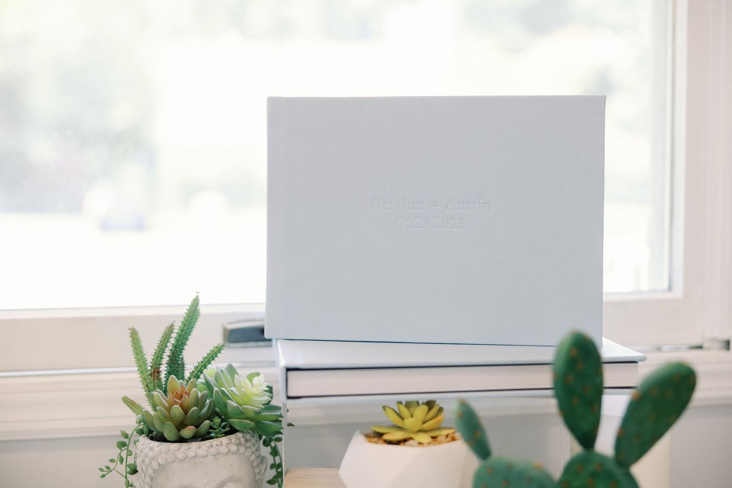 White wedding photo album displayed on a windowsill surrounded by small green succulent plants.