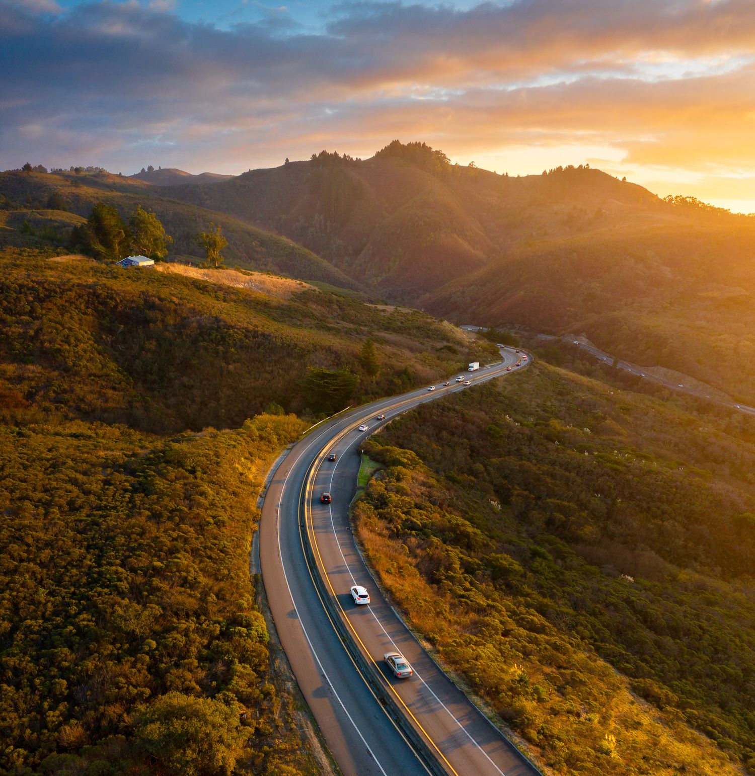 Scenic winding mountain road with cars driving through golden hills during dramatic sunset in California.