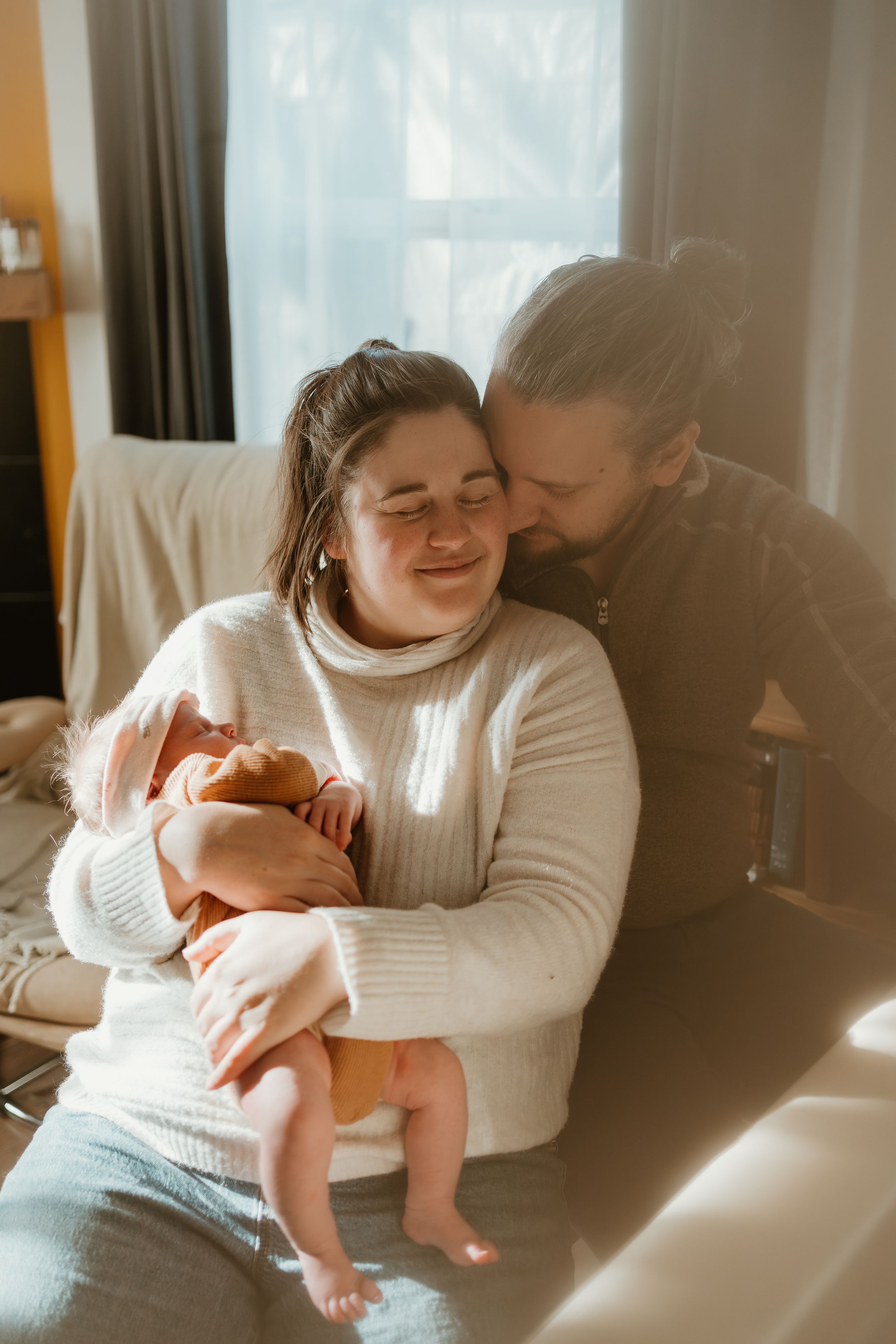 Sunlit indoor family moment with newborn baby in white and brown clothing.