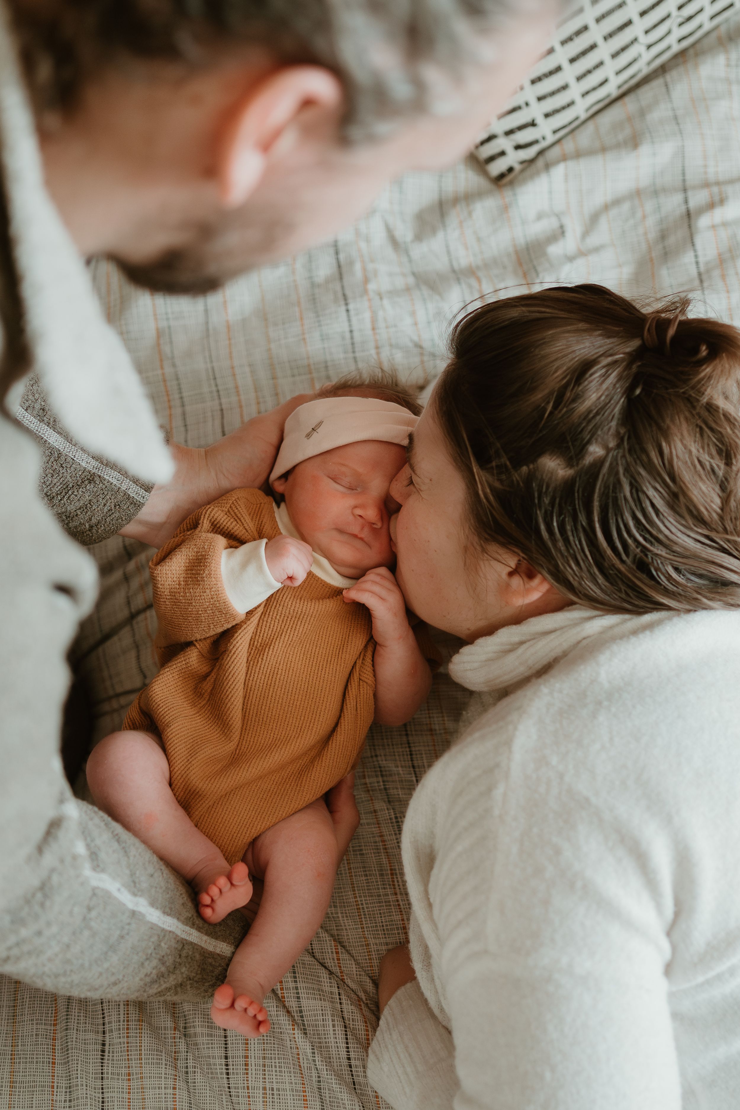 Gentle family cuddles with newborn baby wearing brown clothing on patterned bedding.